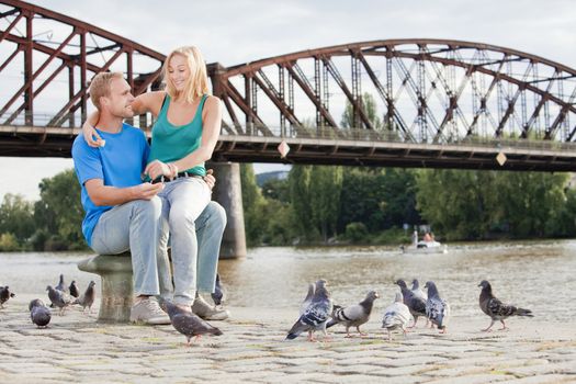 young happy couple sitting by the riverside, feeding birds