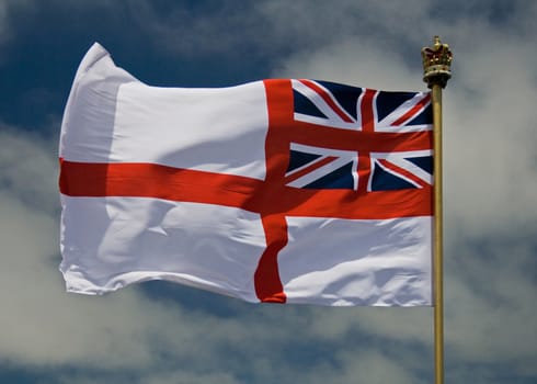 A photo of the British Royal Naval Flag, flying against a blue sky with some light clouds, and a crown on the top of the flag pole