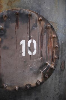 A close-up of a dark, stained release valve on a large metal tank with large bolts and handle. The number 10 overprinted in white.