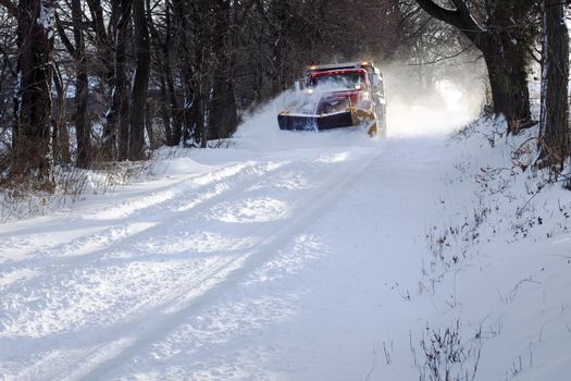 A snowplow truck removing snow from a tree lined rural road on a cold winter day.