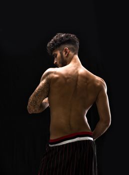 Young athletic man with tattoo posing isolated over black background, back view