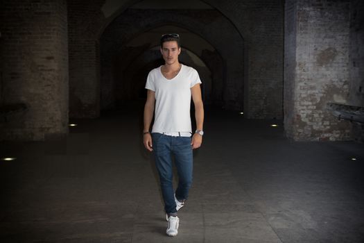 Handsome young man in old building walking inside gallery, looking at camera, full figure shot