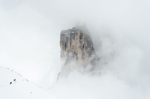 4 people in the lower left corner hiking through the snow in winter around the Tre Cime di Lavaredo, Drei Zinnen, Dolomites mountain peaks partially covered in the clouds.