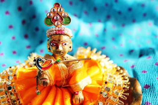 Brass idol of baby lord Krishna, a Hindu god, dressed in bright yellow clothing, against a polka dotted aqua colored cloth background, sporting his legendary musical flute. Ideal for Hindu religious use and festivals - like Janamashtami, Gokul ashtami. 
