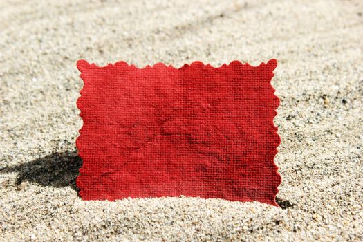 A blank red handmade paper note card with blank space for message standing in sunlit sand. Ideal for beach holiday theme. 
