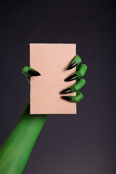 Green monster hand with black nails holding blank piece of cardboard, Halloween theme  