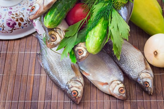 On a table on a chopping board there are a fish, cucumbers, tomatoes, onions, spices and parsley greens.