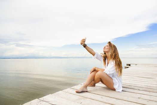 Portrait of beautiful young woman sitting on a pier and taking a self portrait with her smart phone