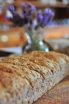 A rustic image of home baked bread on a wooden cutting board with bread crumbs all round. A small vase with lavender blooms blurred in the background.