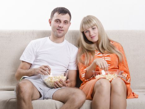 Couple sits on the couch. Each lap Cup of popcorn. Couple watching TV, eating popcorn and smiling.