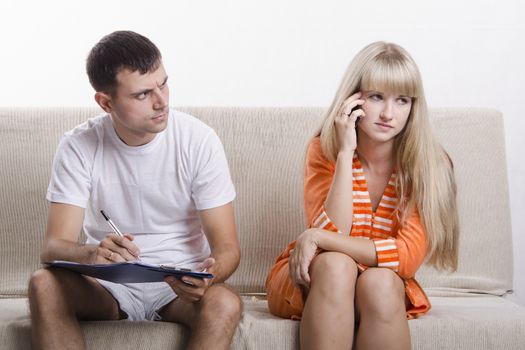Young couple sitting on the couch. Girl on the phone, the guy sitting with the directory and writes a note of the telephone conversation. He is very surprised to hear, girl concerned. The pair dressed home