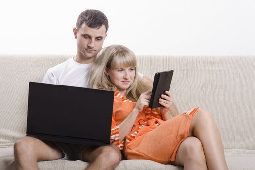 A boy and a girl sitting on the couch. A guy sitting with a laptop, a girl with a tablet. The girl clung to the guy. Both smiling,