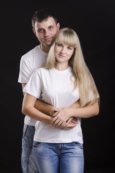 A half-length portrait of a young couple. Guy hugging a girl back. Studio, black background.