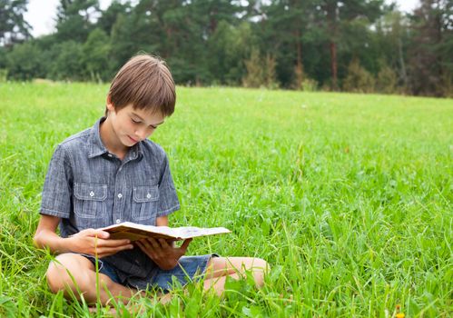 Boy sitting in grass reading a book in a summer field