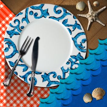 Empty plate with fork and knife, red and white checkered tablecloth, seashells, blue waves and starfish. Table set for a seafood menu