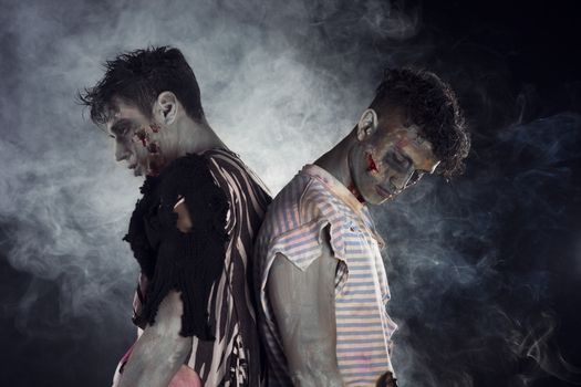 Two male zombies back to back on black smoky background, looking down and away