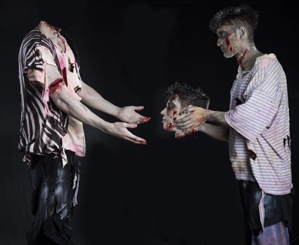 Two male zombies, one giving the head to the decapitated one's body, standing on black background