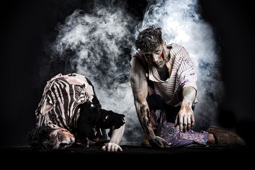 Two male zombies crawling on their knees, on black smoky background, looking at camera. Halloween theme