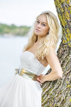 Blond young woman leaning on a tree at the edge of a lake