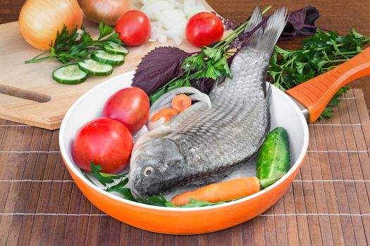 On a table on a chopping board there are a fish, cucumbers, tomatoes, onions, spices and parsley greens.