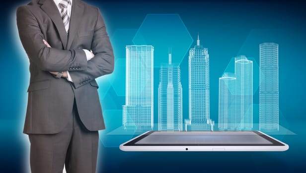 Businessman in suit stands with his arms crossed. Glowing wire-frame buildings on screen tablet pc as backdrop