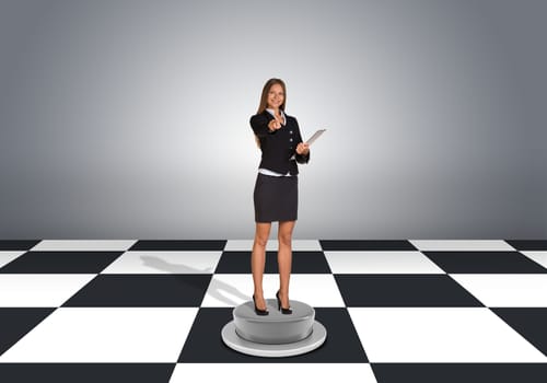 Beautiful businesswoman holding folder and pointing finger at camera. Floor with checkerboard texture and gray wall