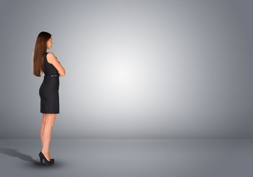 Businesswoman in dress standing in an empty gray room. Rear view. Hands crossed on his chest