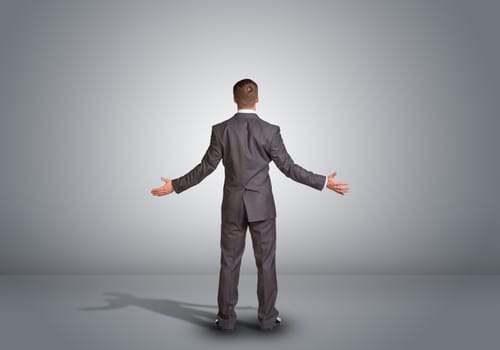 Businessman in suit standing in an empty gray room. Rear view. Spread your arms to side