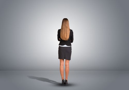 Businesswoman in suit standing in an empty gray room. Rear view. Hands crossed on his chest