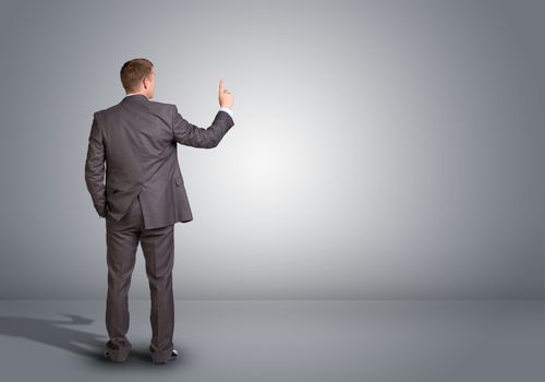 Businessman in suit standing in an empty gray room. Rear view.  Finger pointing up