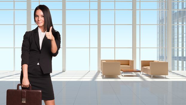 Businesswoman holding briefcase and showing thumb-up. Large window in office building as backdrop