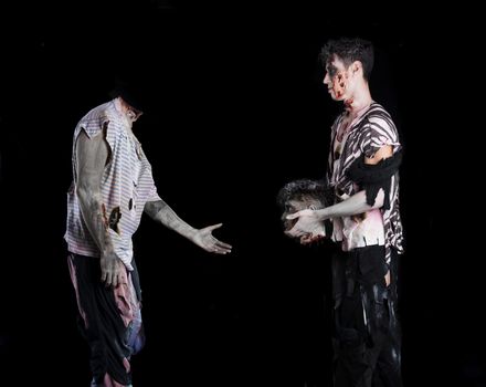 Two male zombies, one giving the head to the decapitated one's body, standing on black background