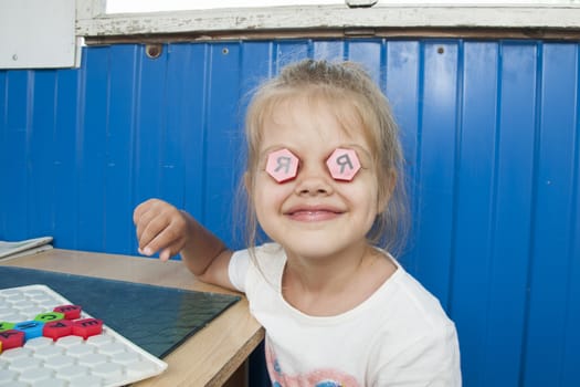 Girl sitting on the veranda at the table and playing a Board game with a mosaic with letters on it. The girl put this in the eyes of the two mosaic with the letters "I".