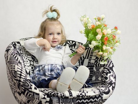 A little girl sitting on a chair with a bouquet of artificial flowers. Palgem points to itself. Smiling. Studio, white background