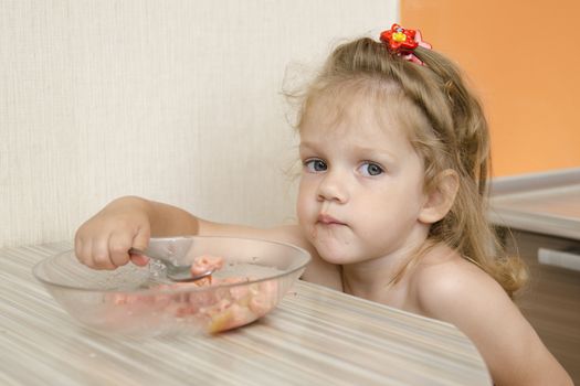 The child sits in the kitchen and eats porridge at the table. The child is distracted and with views of the inquiring looks into the frame.