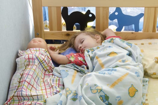 Two-year-old girl sleeping in bed under the covers. Next doll is also covered with a blanket. Children's room.