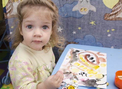 Two-year-old girl is painting at table in children's room. Landscape page with scribbles and smileys. girl surprised looks into frame.