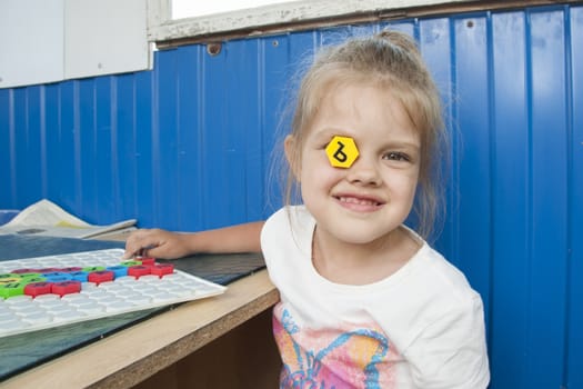 Girl sitting on the veranda at the table and playing a Board game with a mosaic with letters on it. The girl put this in the right eye with the letter of the mosaic