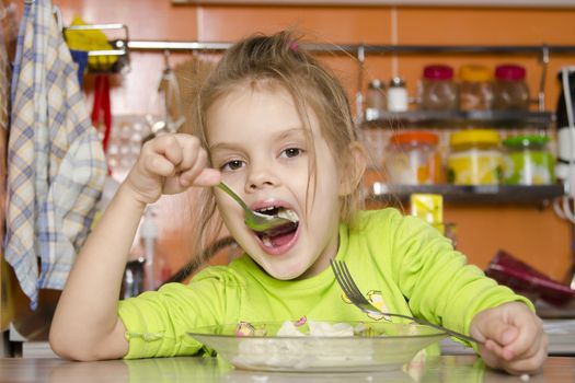 A four year old girl eats sitting at the table in the kitchen. In one arm and the girls spoon in the other fork. Girl eat with a spoon
