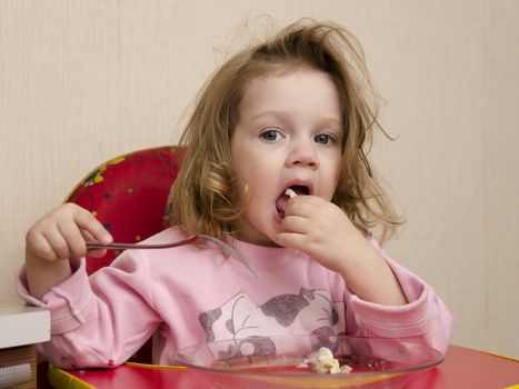 Two-year-old girl eats sitting at the table in the kitchen. Girl eats with a fork.