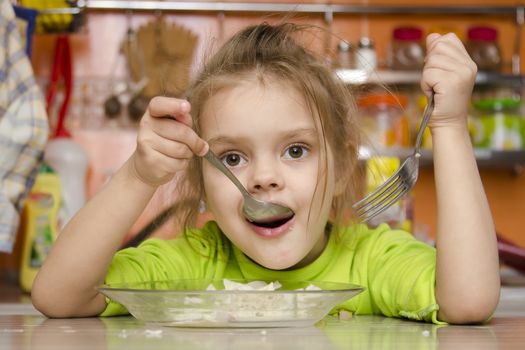 A four year old girl eats sitting at the table in the kitchen. In one arm and the girls spoon in the other fork. Girl eat with a spoon