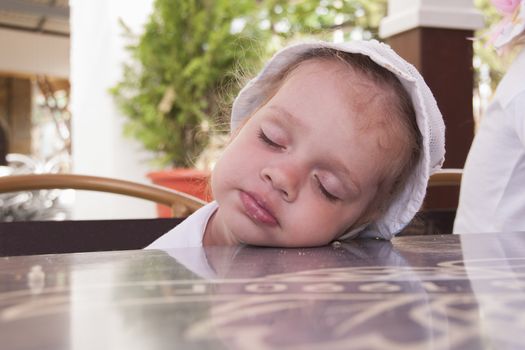 Two-year-old girl eating in the street cafes fell asleep sitting on a chair and resting his head on the table.