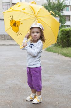 Merry little girl stands with the yellow umbrella on an asphalt road on the street