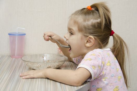 Girl eating porridge sitting at the table. The profile view. Home furnishings