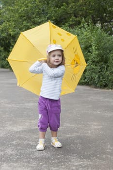 Merry little girl stands with the yellow umbrella on an asphalt road on the street