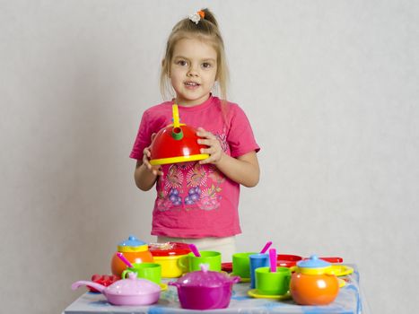 Girl plays child kitchen utensils. She picked up the teapot and looks in the frame. A light background, a Studio.