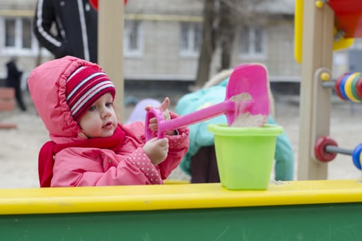 The little girl pours sand in the bucket, while in the sandbox. In early spring. Girl dressed in warm clothes