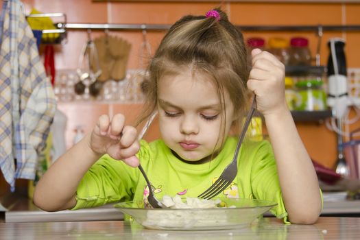 A four year old girl eats sitting at the table in the kitchen. In one arm and the girls spoon in the other fork. Girl eat with a spoon