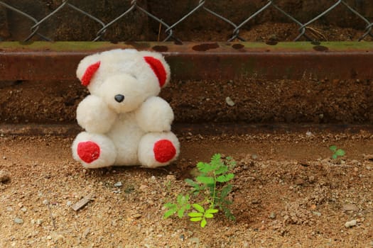 The doll is placed at the fence near the green plant.