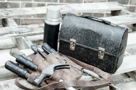 Construction worker's lunch break with lunch pail, beverage container, tool belt, hammer, screwdrivers, and wrenches on wooden pallet with brick wall in background. Image has been moderately desaturated. 